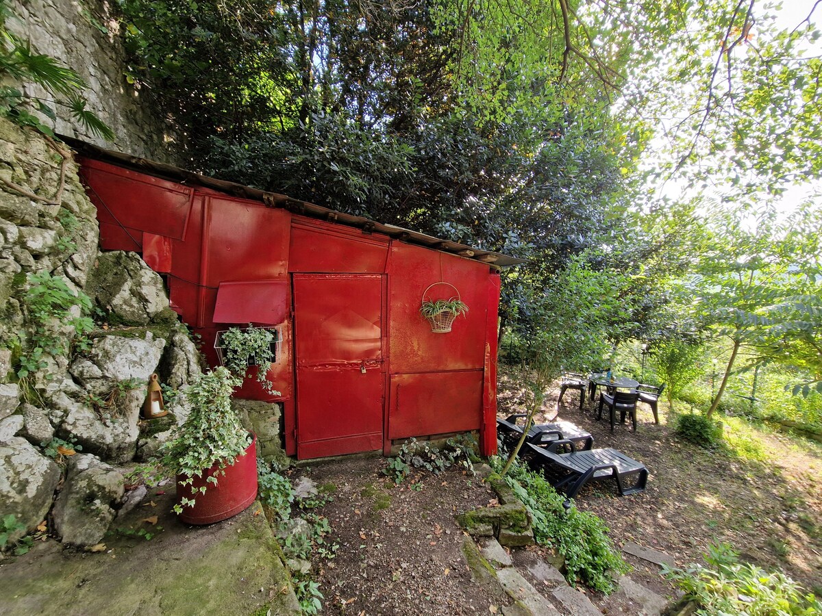 A vibrant red structure is set against a backdrop of lush green foliage. Stone steps lead down to a small outdoor seating area with a table and four chairs, surrounded by trees and natural surroundings.