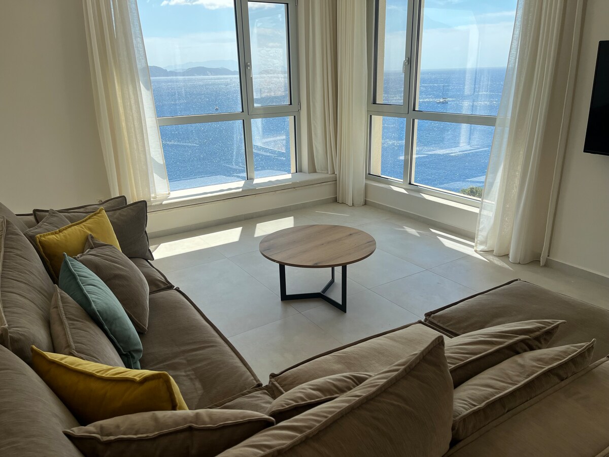 A spacious living area features large windows that provide uninterrupted views of the Ionian Sea. A comfortable sectional sofa, adorned with colorful cushions, is positioned around a round coffee table, inviting relaxation and enjoyment of the scenic backdrop.