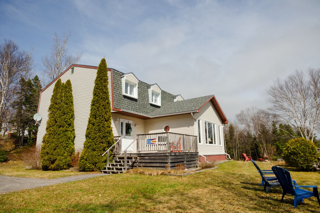 A two-story house is set within a spacious landscaped yard. The exterior features light siding and a sloped roof. Tall conifer trees frame the entrance, which is accessible by a wooden staircase. Deck chairs are positioned on a small patio, inviting outdoor relaxation.