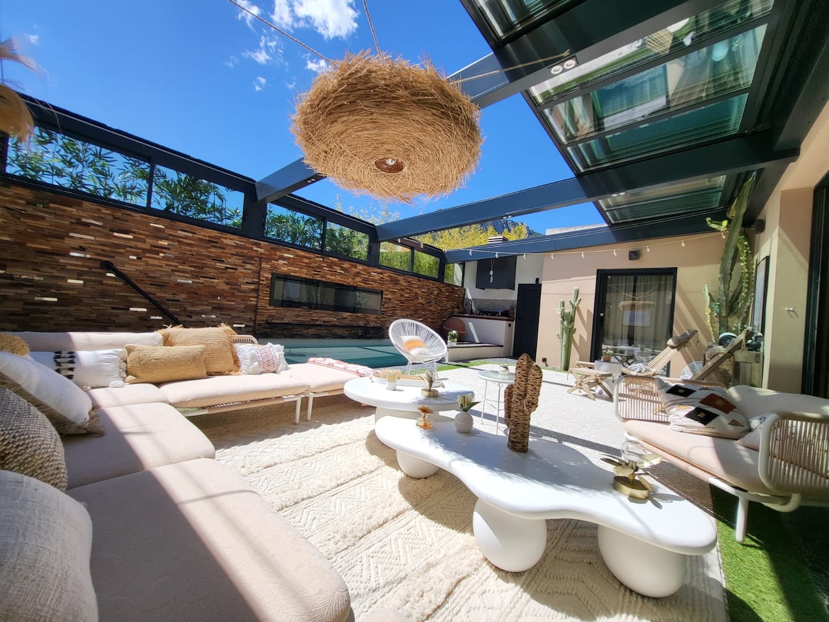 An inviting outdoor lounging area is showcased, featuring a spacious sectional sofa with neutral upholstery. A large hanging light fixture adds a unique touch, while a heated pool is visible in the background, surrounded by greenery. Natural light streams through the glass roof.