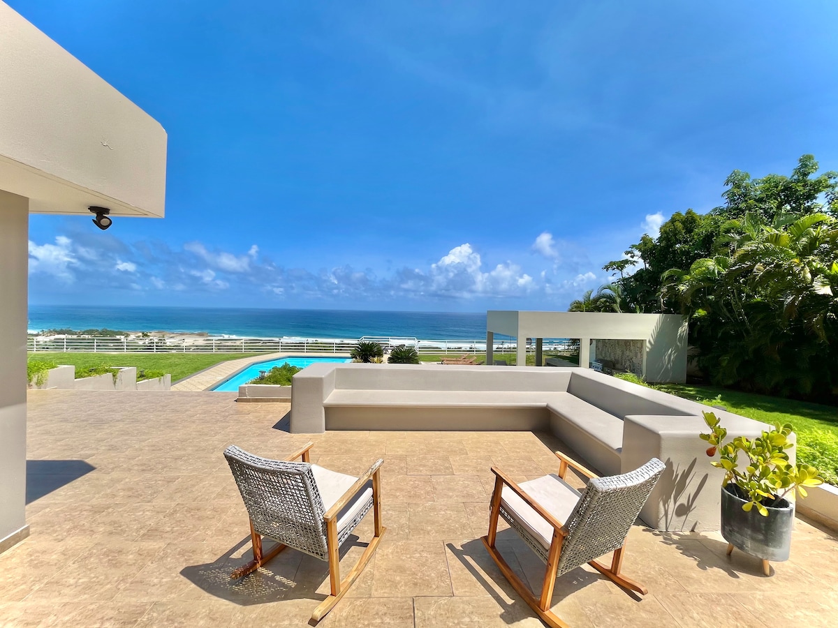 An outdoor seating area is presented with two wicker chairs placed on a tiled patio, overlooking a swimming pool and expansive ocean views. Lush greenery frames the space, enhancing the coastal ambiance under a clear blue sky.
