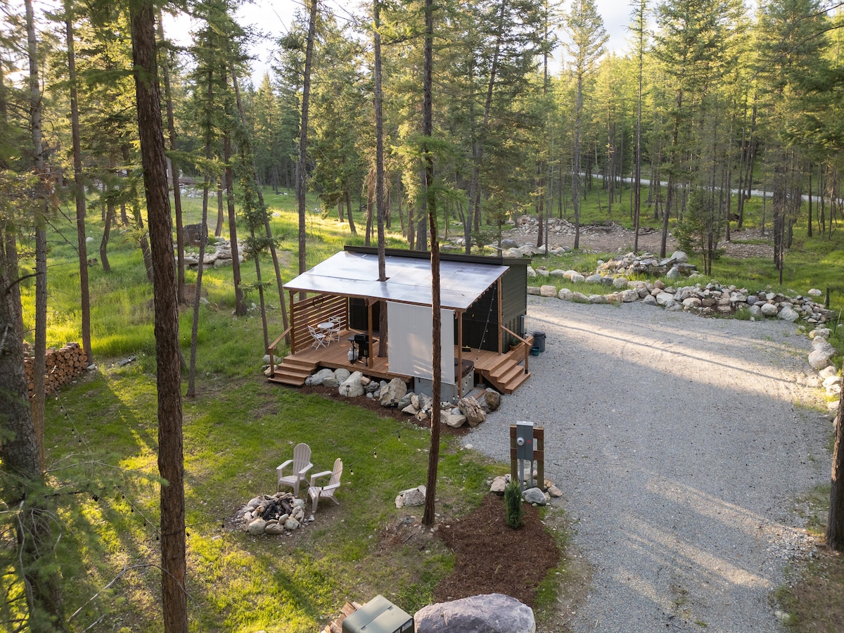 A tiny home nestled among tall evergreens is surrounded by a gravel pathway and natural landscaping. A wooden porch features seating areas, while a woodpile is neatly stacked nearby. The setting emphasizes the peacefulness of the wooded environment, inviting relaxation in nature.