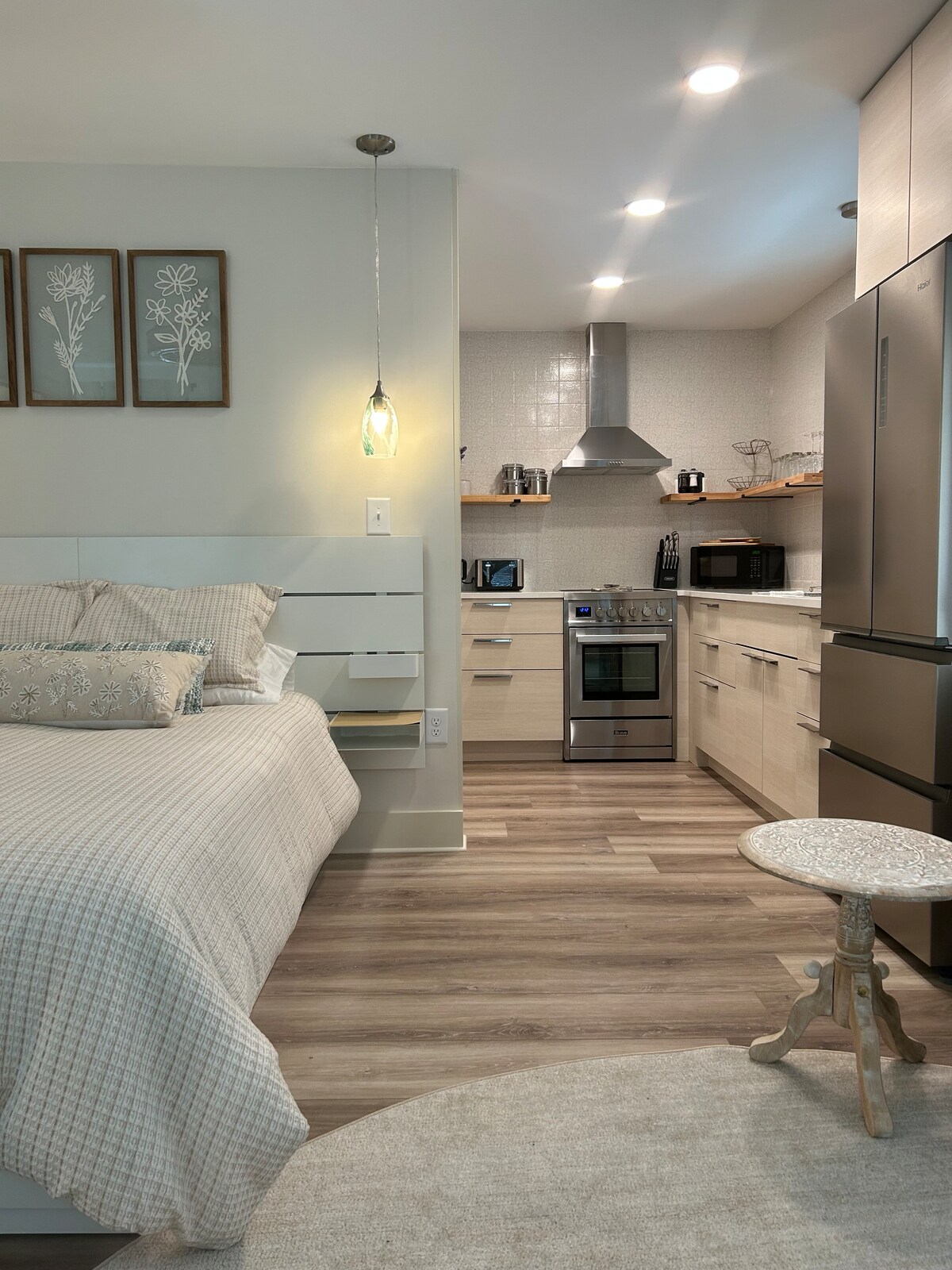 A studio apartment is depicted, featuring a queen bed dressed in light-colored bedding. The full kitchen includes modern appliances and stylish cabinetry. A small, round stool is positioned near the bed, and the space is illuminated by soft overhead lights.
