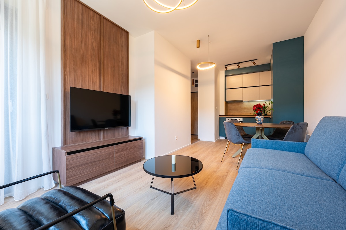 A modern living space is showcased, featuring a comfortable blue sofa and a sleek black coffee table. A large flat-screen TV is mounted on a wood-paneled wall. In the background, a dining area is visible with a round table and chairs, complemented by a stylish kitchen.