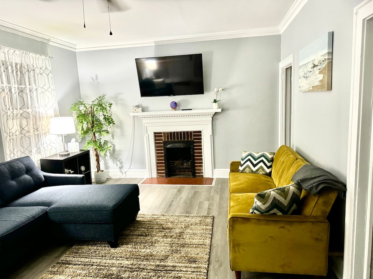 A comfortable living area features a sectional sofa in blue and a yellow accent chair, providing a cozy seating arrangement. A television is mounted above a decorative fireplace, while a light rug adds warmth to the space. Natural light filters through a window with sheer curtains.