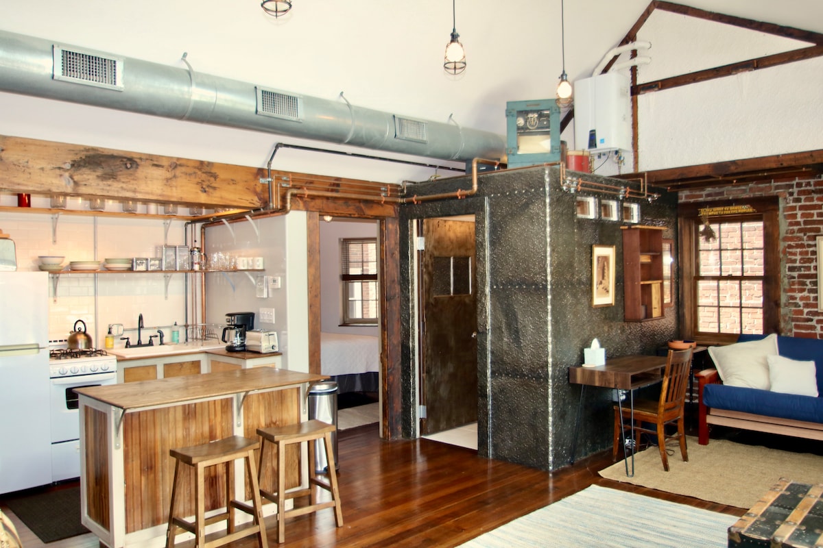 An open-concept kitchen and living area features hardwood floors and exposed wooden beams. A modern kitchen with a countertop and stools is complemented by a rustic dining table and chairs. A cozy seating area showcases a blue couch and large windows allowing natural light.