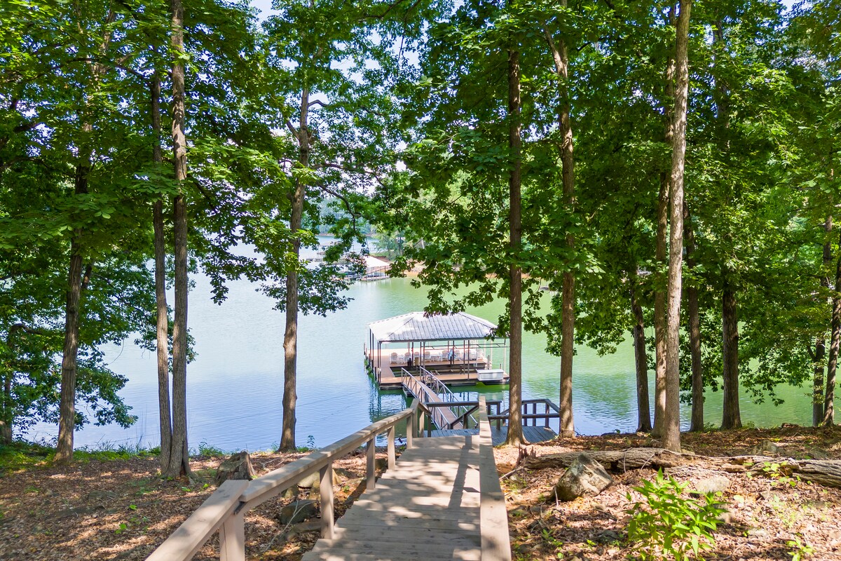 A wooden path leads through a wooded area towards a covered boat dock. The dock is positioned on calm lake waters, surrounded by greenery, providing a serene view. Sunlight filters through the trees, casting dappled shadows on the ground.