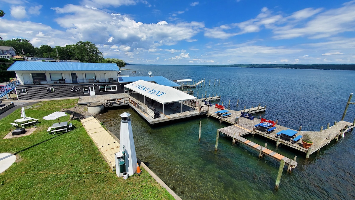 A waterfront view showcases a restaurant with outdoor seating and a large deck extending over the calm waters. Docking spaces are visible alongside the structure. A green lawn leads to the water's edge, complemented by white lounge chairs and umbrellas for guests to relax.