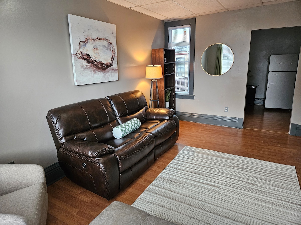 A comfortable living area features a brown leather sofa with a decorative pillow and a side lamp. A round mirror hangs on the wall, reflecting natural light. A textured area rug covers the wooden floor, and a glimpse of a kitchen is visible through an open doorway.