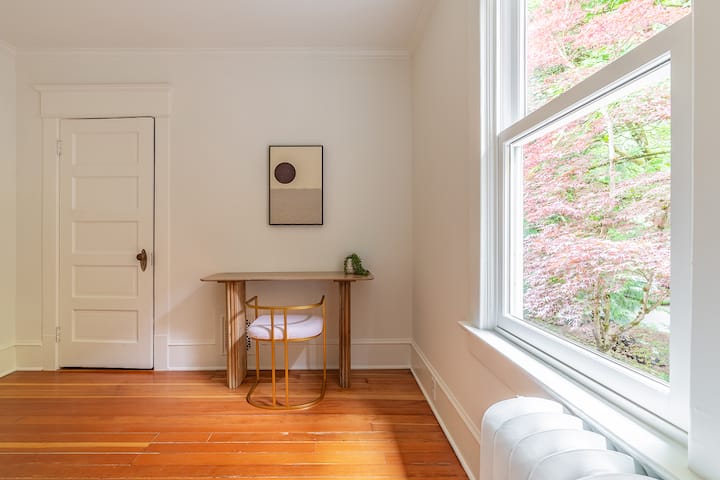 Уютно работно място: осветено от слънцето бюро до големи прозорци, заобиколено от ярка зеленина.