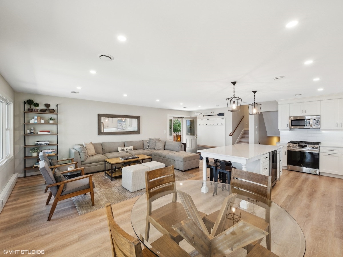 A spacious living area features a large sectional sofa centered around a coffee table. Natural light enters through multiple windows, illuminating the modern open kitchen equipped with stainless steel appliances. A dining table with wooden chairs is positioned nearby, enhancing the inviting layout.