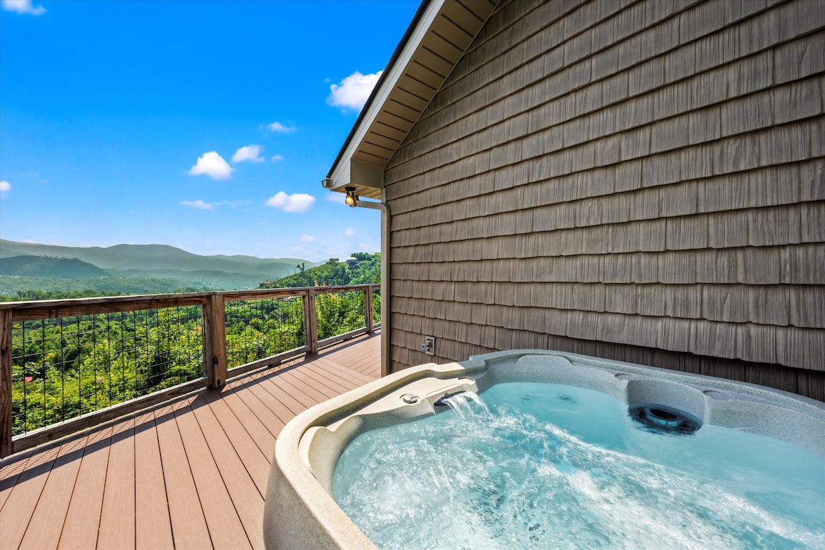 A hot tub is situated on a spacious deck, offering views of the mountains and lush greenery. The wooden railing frames the picturesque landscape beneath a bright blue sky. Clear waters in the tub provide an inviting view for relaxation.