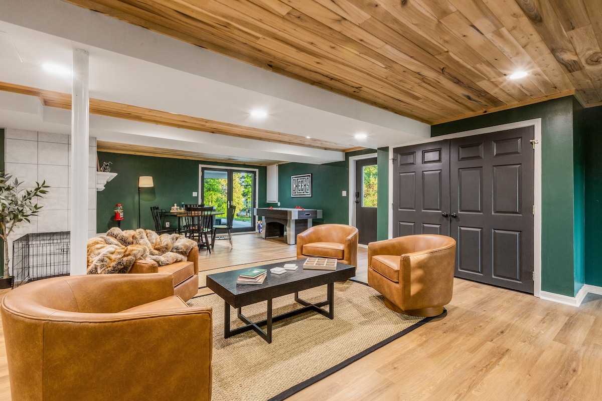 A spacious basement living area is depicted with warm wood paneling on the ceiling and green walls. Two brown leather chairs are positioned around a dark coffee table, set atop a simple area rug. A dining area with a table can be seen in the background, alongside a game table.