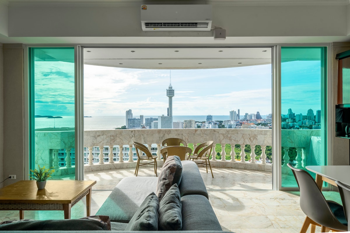 A panoramic view of Pattaya's skyline and coastline is captured through large, sliding glass doors. The spacious living area features a comfortable couch and a dining table, inviting relaxation while overlooking the vibrant cityscape. Natural light floods the space, enhancing the open, airy feel.