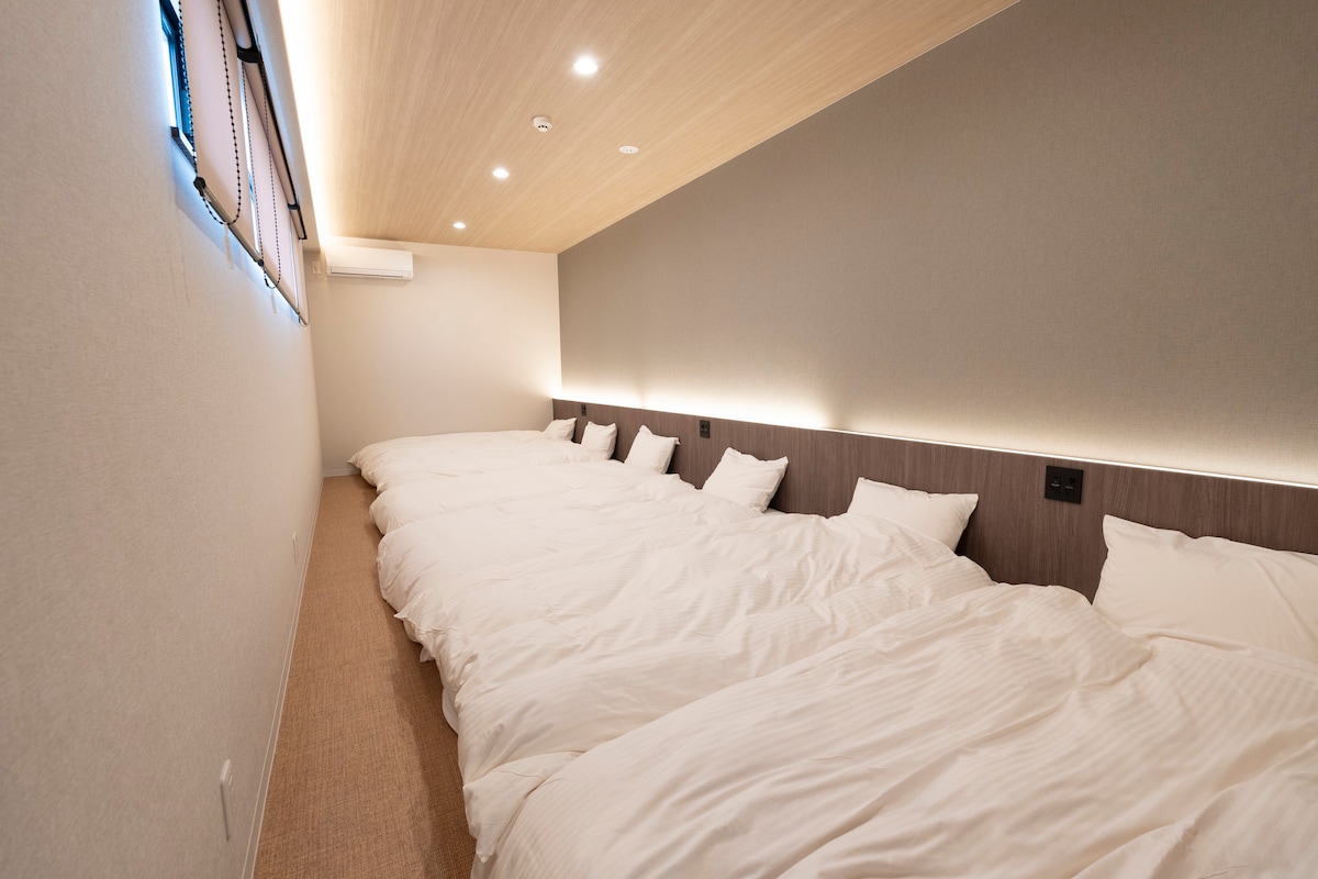 A cozy bedroom is arranged with six spacious mattresses, oriented against a softly lit wall. Warm ambient lighting highlights the clean, minimalist décor, while large windows allow natural light to filter in.
