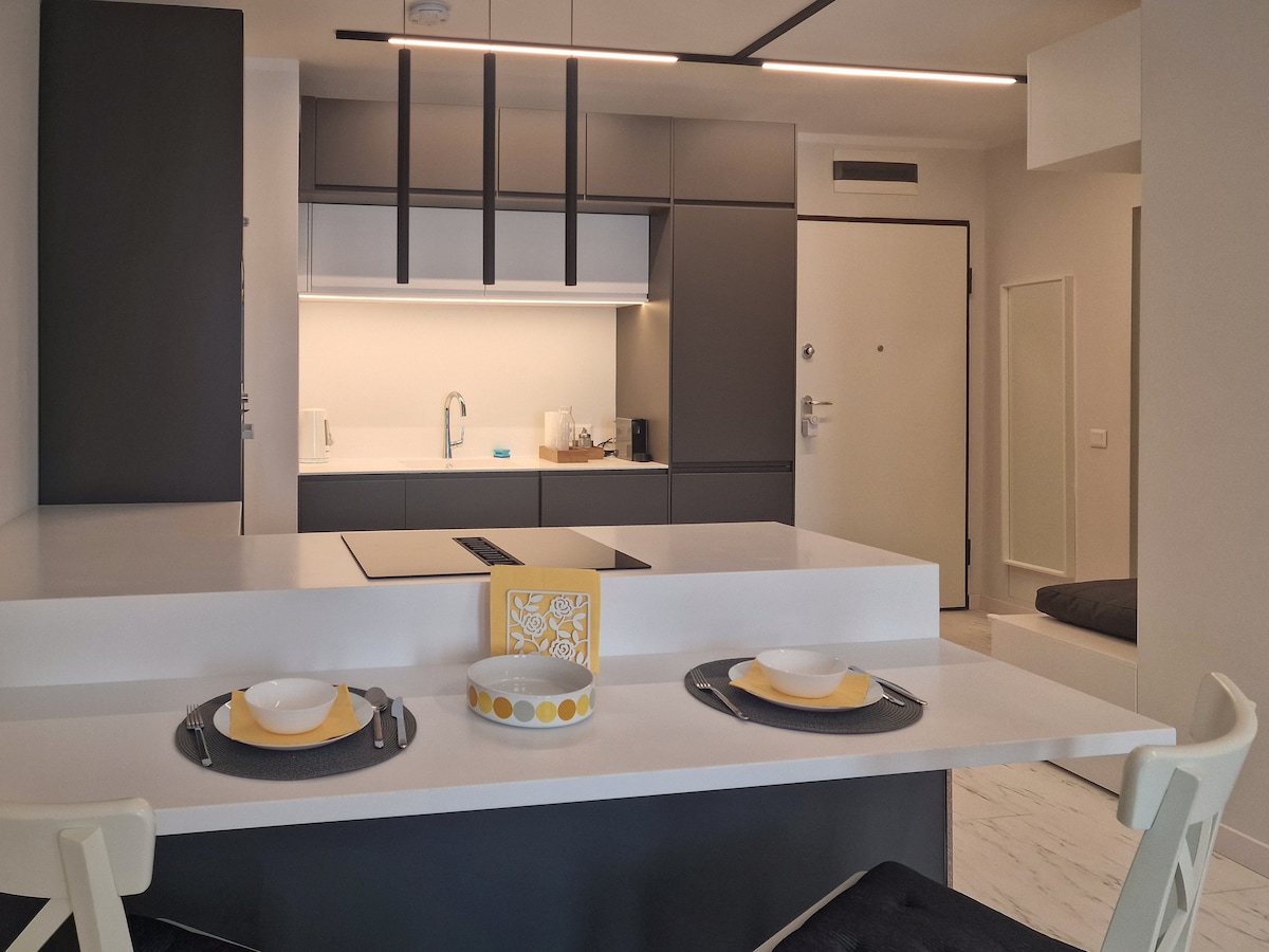 The sleek kitchen features modern cabinetry and integrated appliances. A spacious island serves as a prep area and divides the dining space, which includes two place settings with minimalist tableware. The room is illuminated by soft overhead lighting, enhancing the contemporary design.