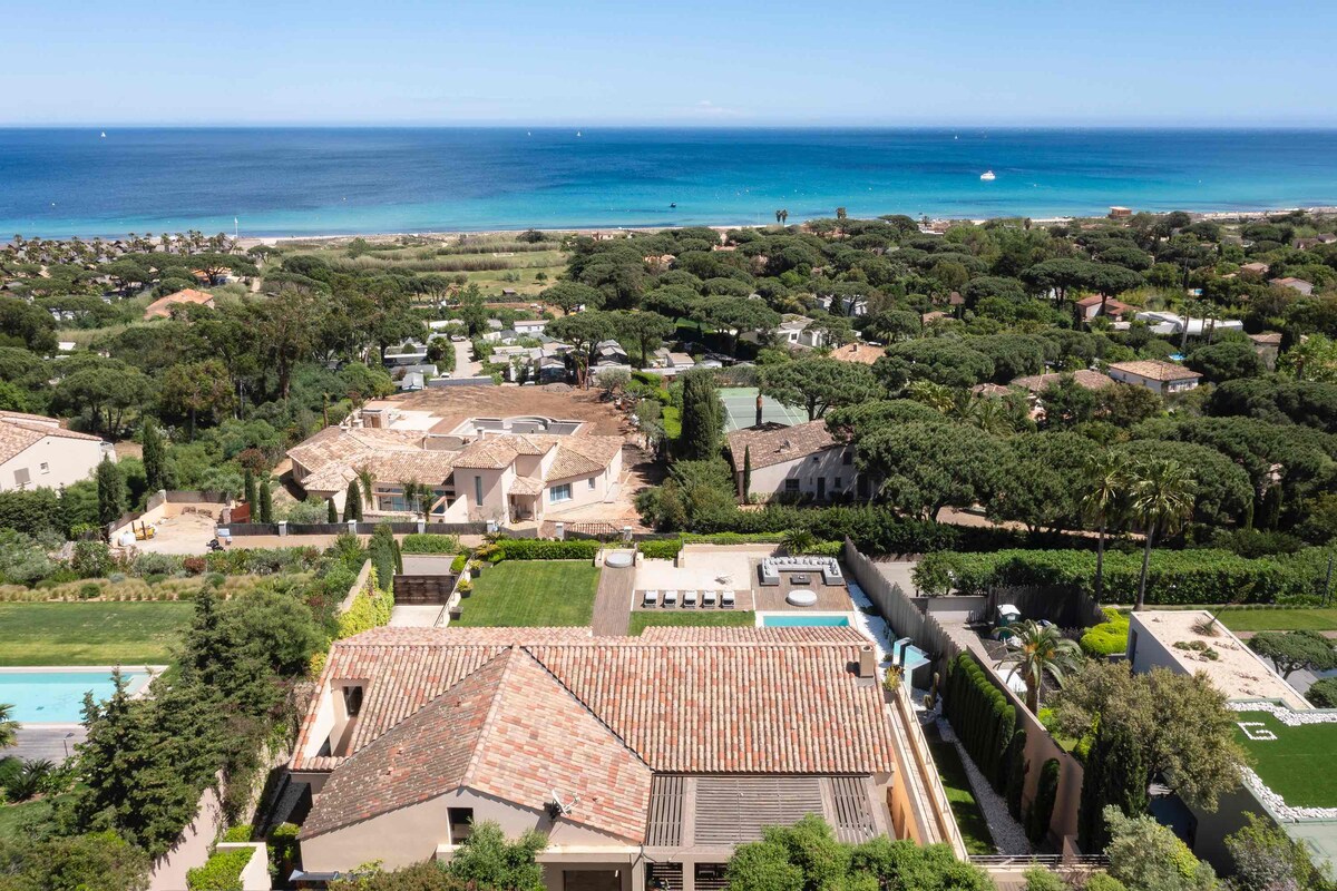 A panoramic view captures the expansive landscape of lush greenery leading to the ocean. The terracotta rooftops of the house contrast with the vibrant blue sea. The inclusion of neighboring properties and a distant beach enhances the tranquil setting.