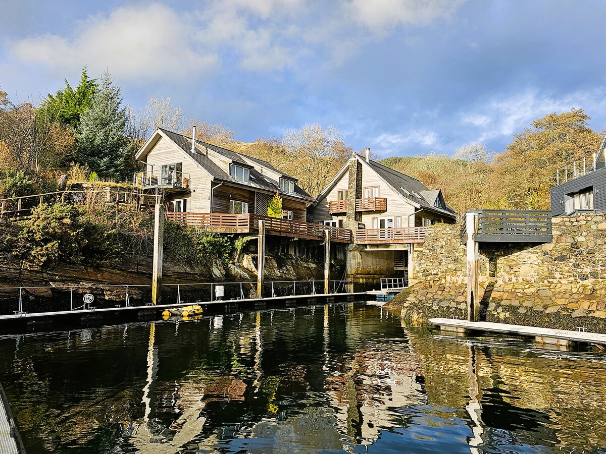 The image showcases a wooden structure nestled against a rocky landscape, featuring spacious decks overlooking calm waters. The building's exterior blends with the natural surroundings, while reflections are visible on the water's surface. Green foliage and autumnal trees frame the scene.