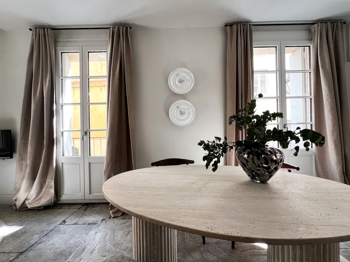A dining area is presented with a round travertine table at its center, accompanied by wooden chairs. Tall windows are adorned with light drapes on either side, allowing natural light to enhance the space. Decorative wall pieces above the table complete the elegant décor.