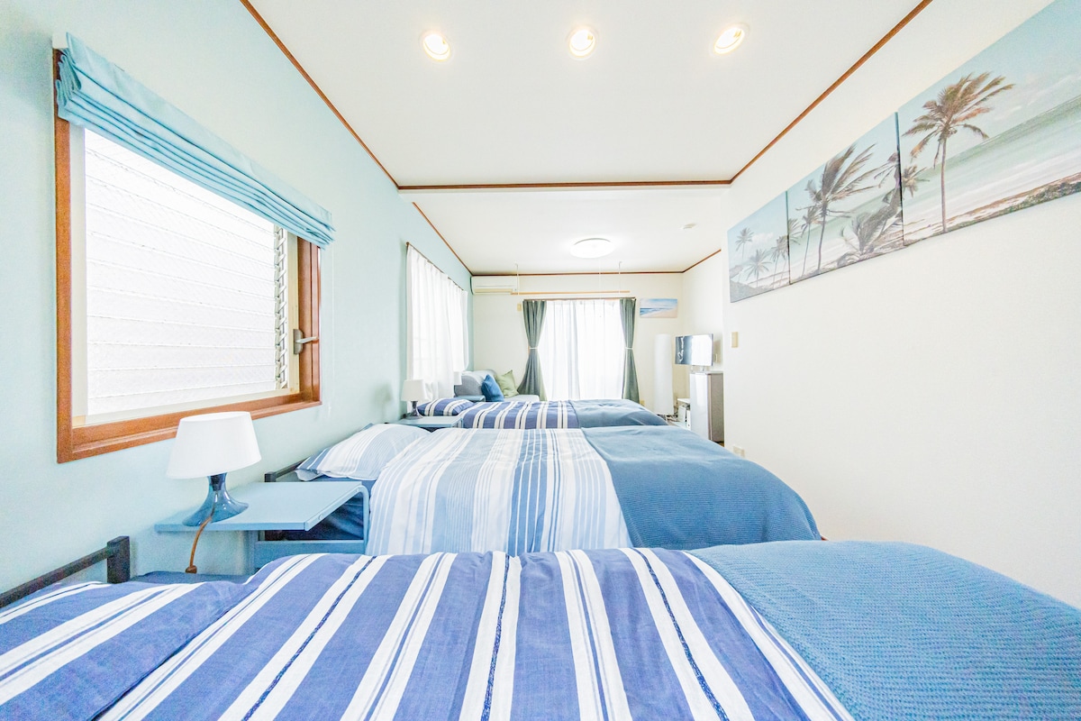 A bright and airy bedroom contains multiple beds with blue and white striped bedding. Natural light fills the space through a window adorned with light blue curtains. A small bedside table and a piece of wall art depicting a beach scene enhance the room's relaxed atmosphere.