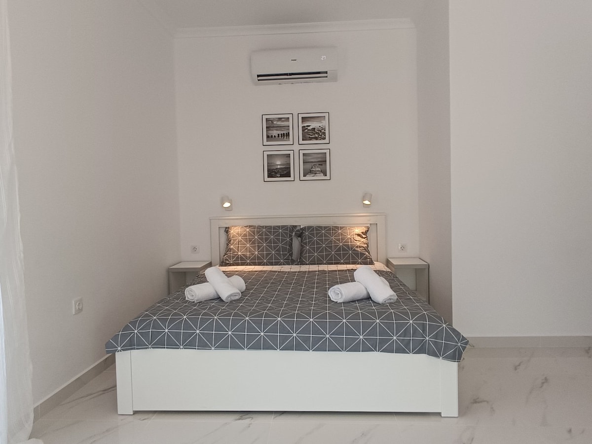 A spacious sleeping area is presented with a double bed featuring a geometric patterned cover. Soft lighting is provided by bedside lamps, highlighting framed photographs on the wall above. Fresh towels are neatly arranged on the bed, adding to the room's inviting ambiance.
