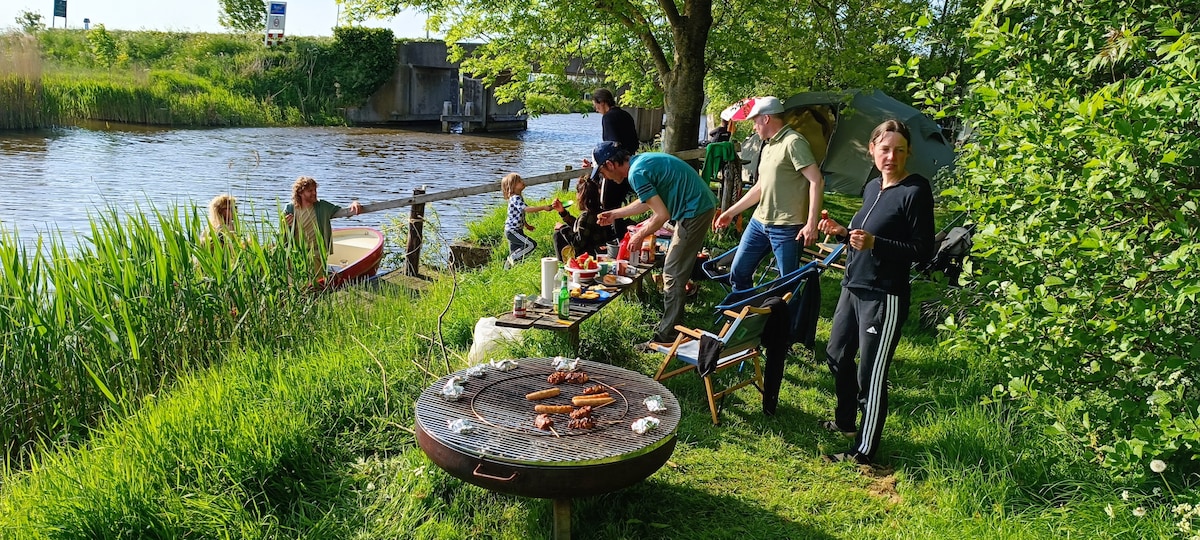 A vibrant outdoor scene captures a group gathered by the water, preparing a barbecue on a circular grill. Lush greenery surrounds them, while a nearby boat floats along the calm water. Relaxation chairs and an umbrella provide comfort for those enjoying the sunny setting.