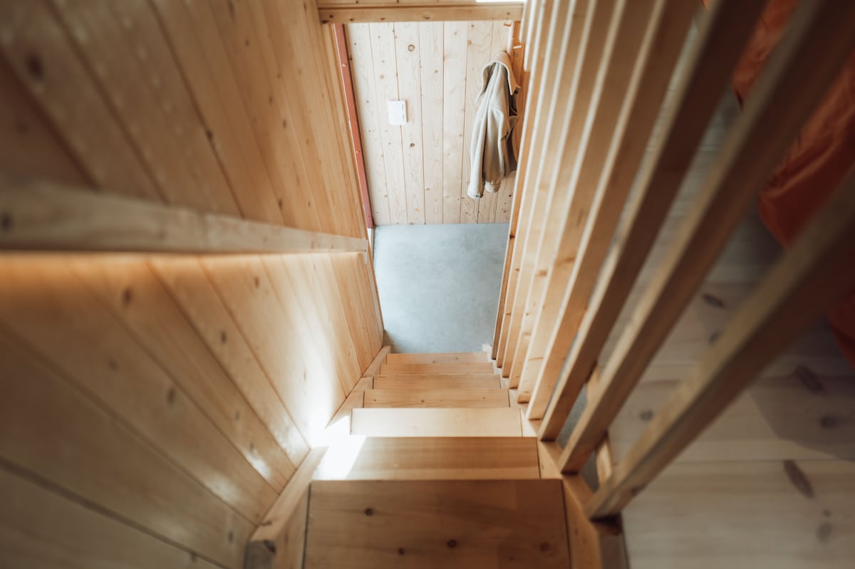 A wooden staircase leads down, framed by vertical wooden railings. Natural light filters in from a doorway at the bottom, highlighting the smooth concrete floor. The environment is tranquil, with a minimalist design emphasizing the warmth of the wood.