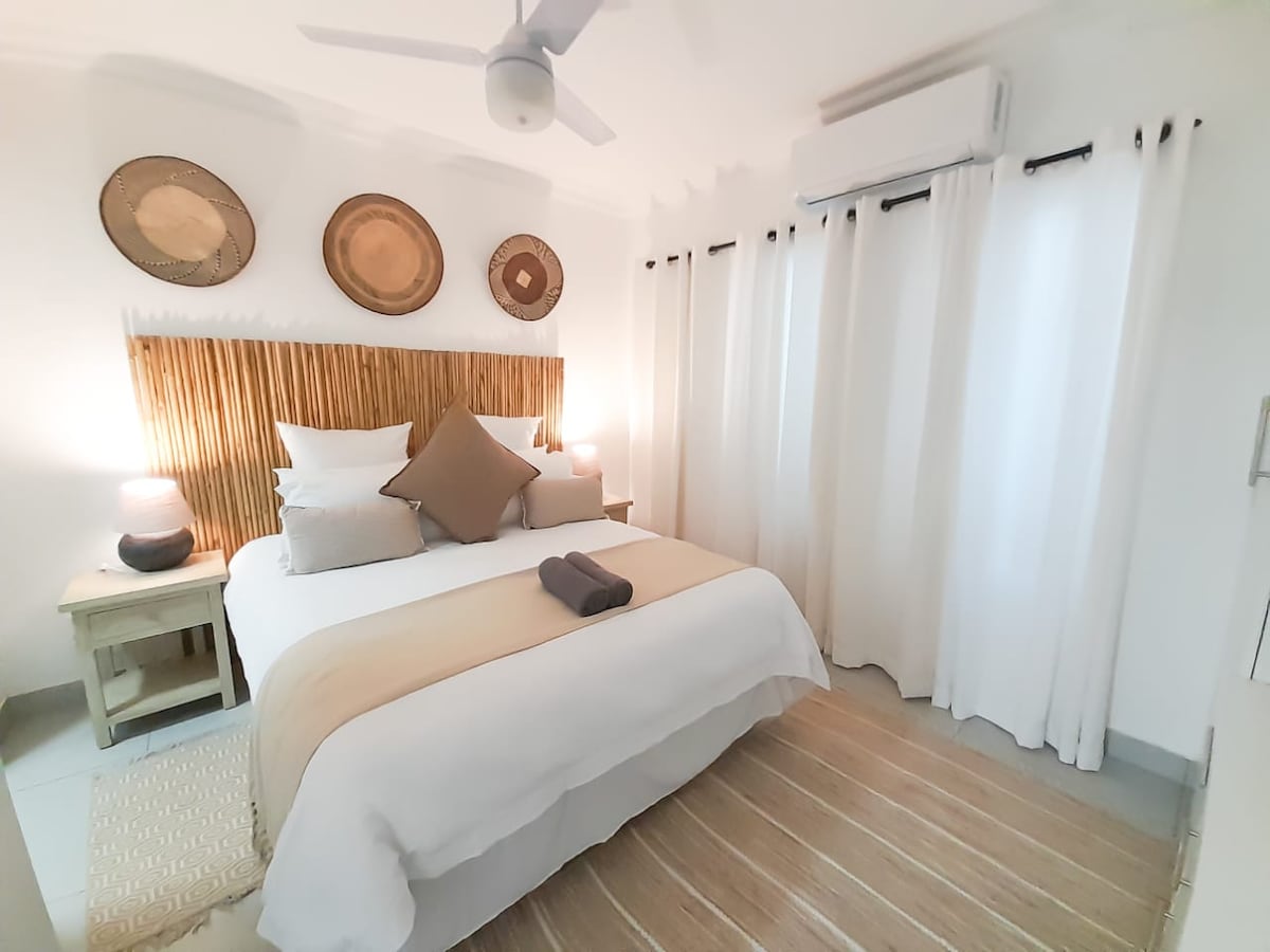 A bright bedroom features a spacious bed adorned with neutral linens and decorative pillows. Natural elements are included with woven wall art, while two bedside lamps provide a soft glow. Light curtains frame the window, and a ceiling fan ensures air circulation.