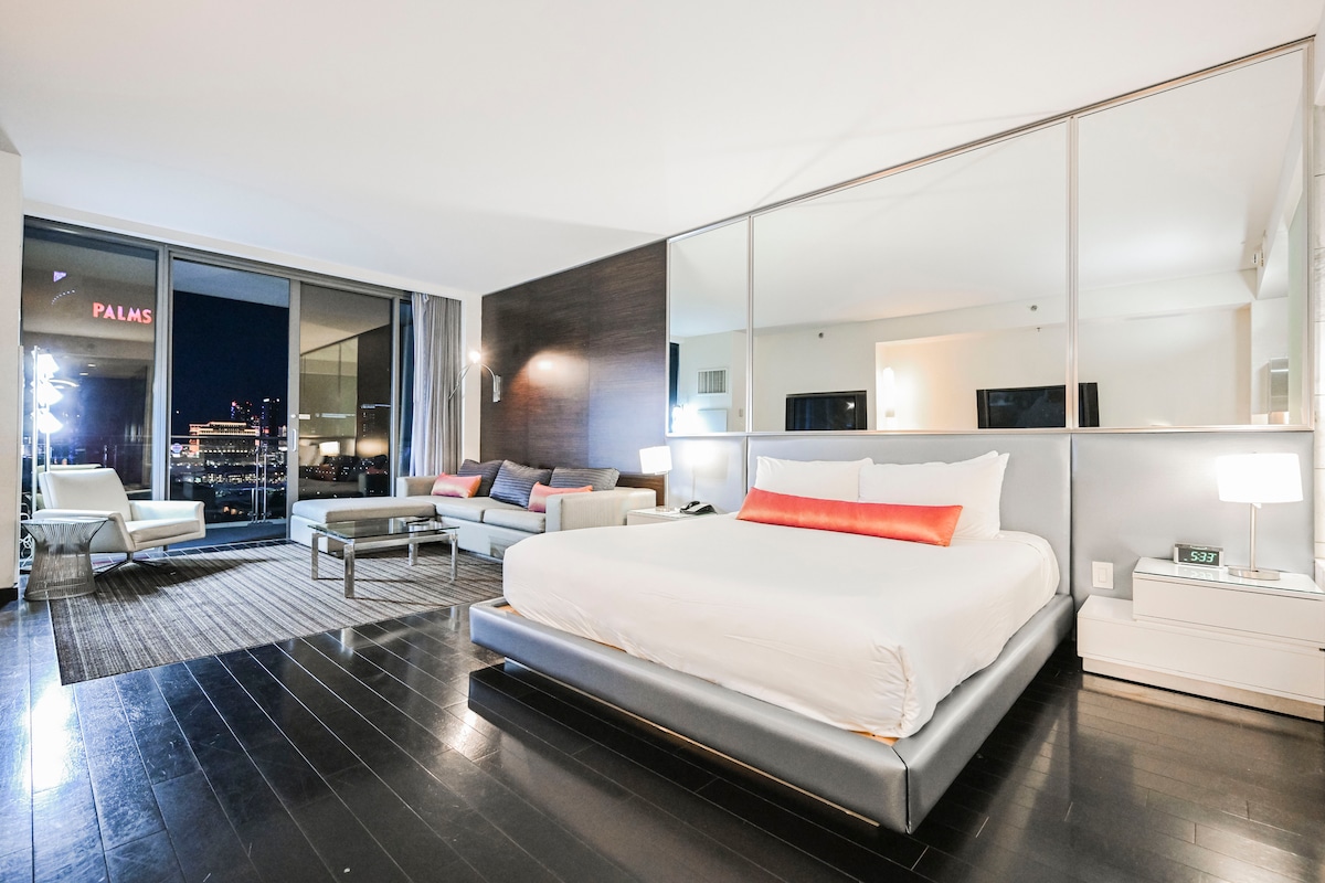 A spacious studio apartment features a king-size bed with a sleek headboard, complemented by a modern sofa and chairs in the living area. Large windows provide views of the Las Vegas Strip, with city lights visible at night. A patterned rug adds texture to the dark wood floor.