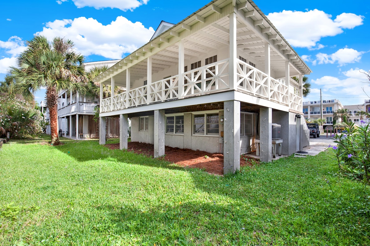 The exterior of a two-story cottage is shown, featuring a large wrap-around porch supported by white pillars. Lush green grass surrounds the base of the structure, with palm trees visible in the background. A clear blue sky adds to the coastal ambiance.