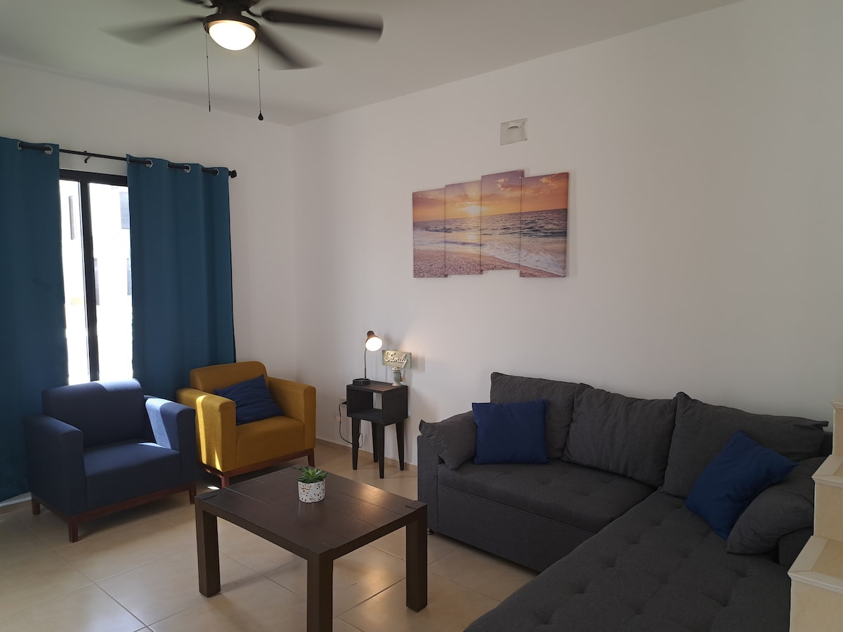 A comfortable living area features a sectional sofa and two armchairs, one in yellow and another in dark blue. A dark wood table sits at the center, accompanied by a potted plant. Light streaming in from a window is filtered by blue curtains, while beach-themed artwork adorns the wall.