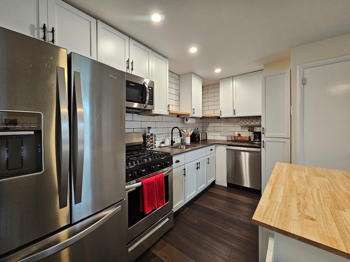 A modern kitchen featuring stainless steel appliances, including a refrigerator and gas stove. White cabinetry complements rustic countertops, while a portable island offers additional countertop space. Natural wood shelves display cooking essentials, creating an efficient and inviting cooking area.