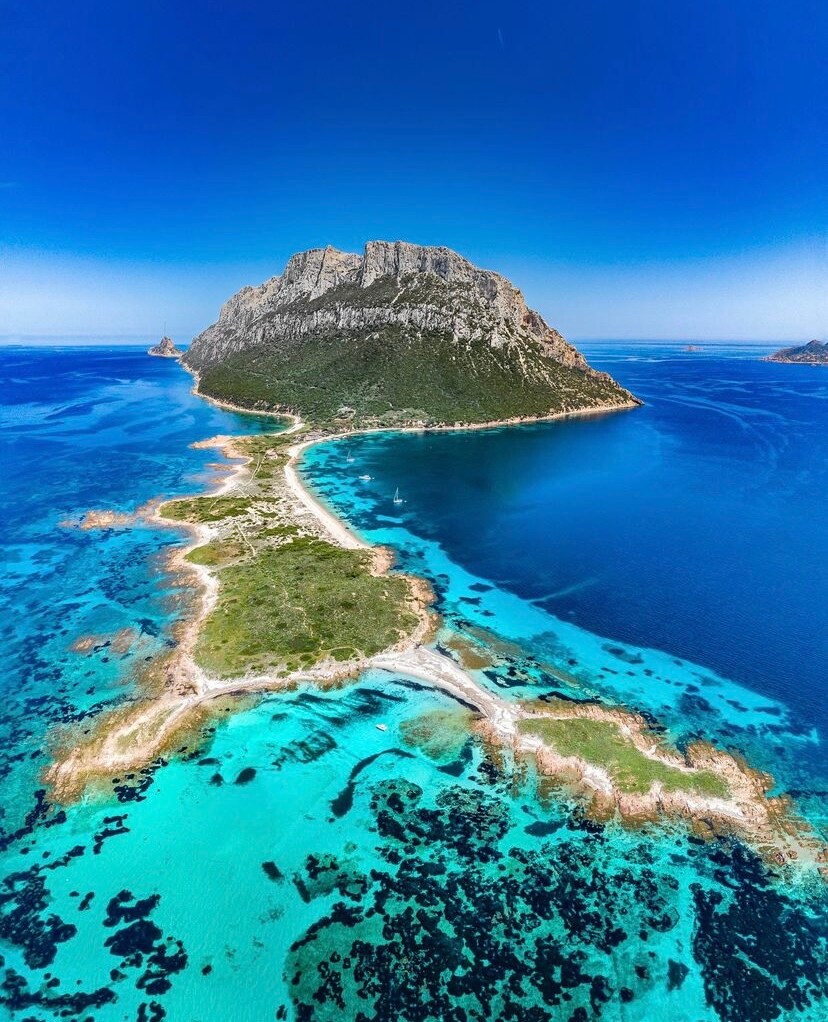 An aerial view captures the stunning Tavolara island, characterized by its prominent rocky peak surrounded by vibrant blue waters. The coastline features a blend of lush greenery and rocky outcrops, while the clear, shallow waters reveal hints of marine life beneath the surface.