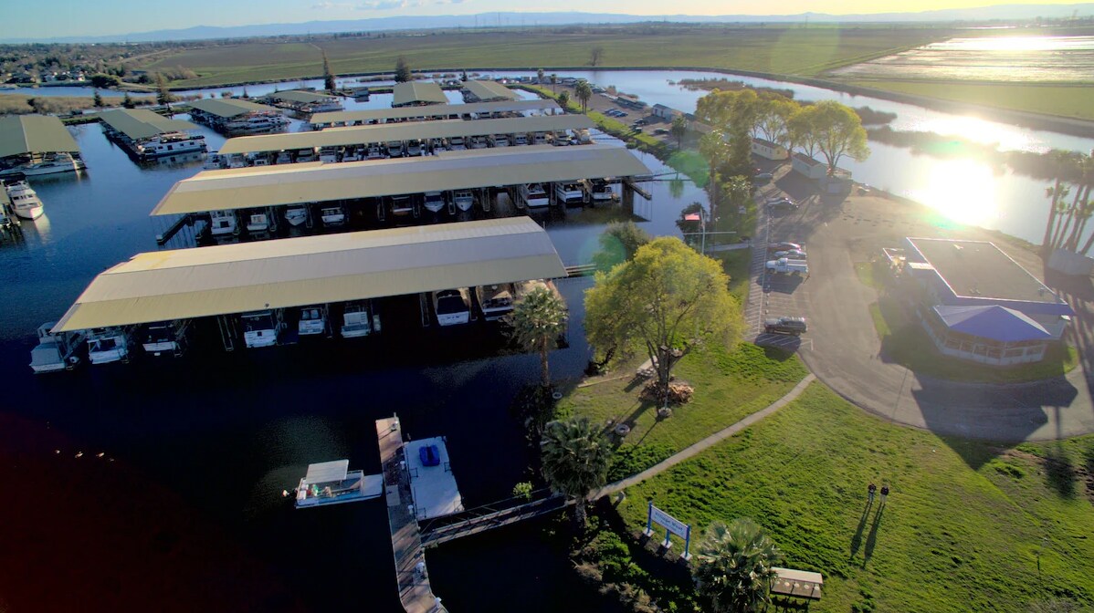 Aerial view captures a marina with covered boat slips reflected in the calm water. A winding path leads to a small parking area and lush green grass, offering glimpses of the surrounding landscape and waterways.
