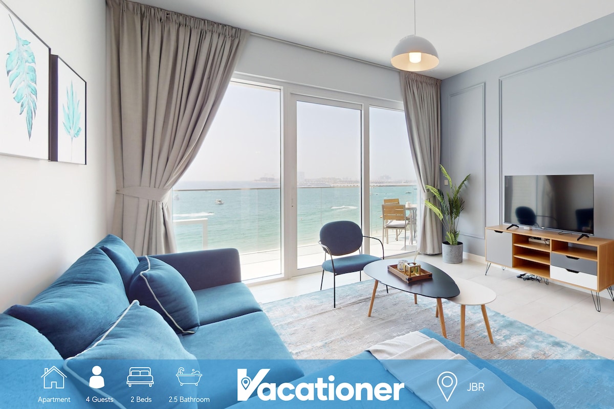 A spacious living area features large windows with views of the sea. A comfortable, L-shaped sofa with plush cushions is complemented by a stylish coffee table and a smart TV. Natural light floods the room, enhancing the serene coastal atmosphere.