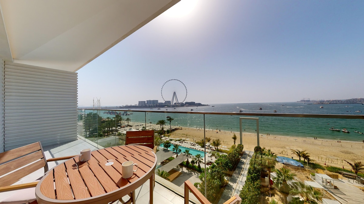 A furnished balcony offers a relaxing view of the beach and sea, with Ain Dubai in the distance. A round dining table with two chairs is positioned beside two cups, inviting outdoor enjoyment of the scenic surroundings. Sunlight streams in, highlighting the bright coastal atmosphere.