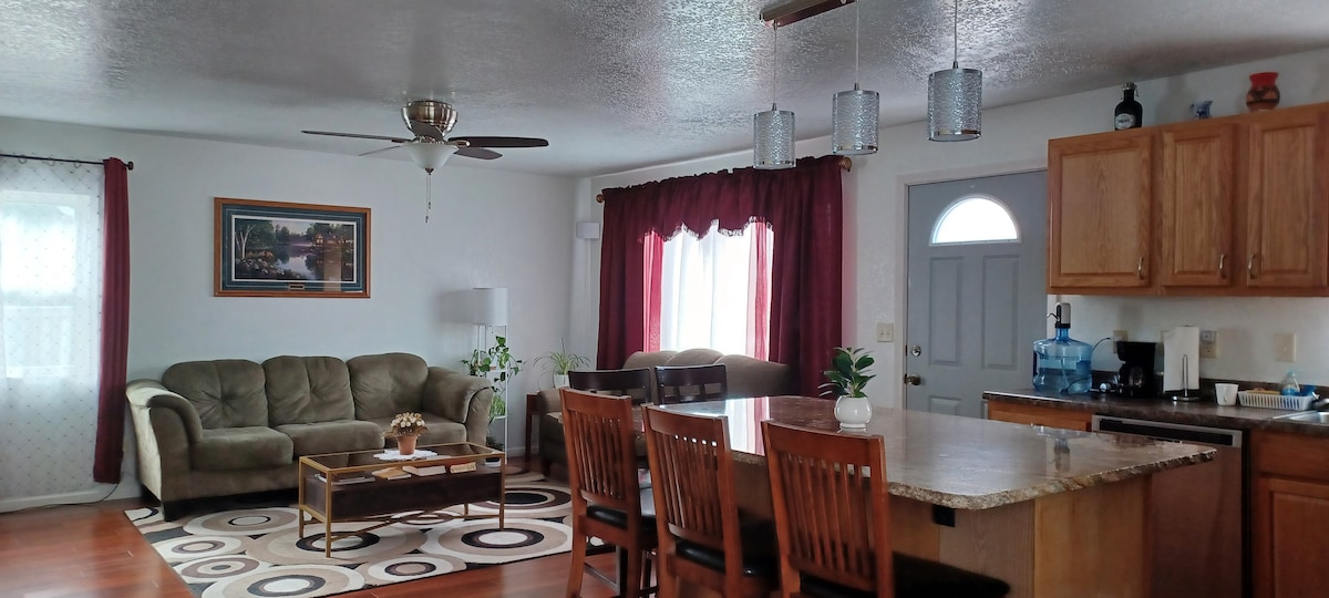 An open living area features a comfortable seating arrangement with a sofa and coffee table, set against a backdrop of natural light from large windows. An inviting kitchen area includes a breakfast bar with seating, modern appliances, and decorative items adorning the countertops.