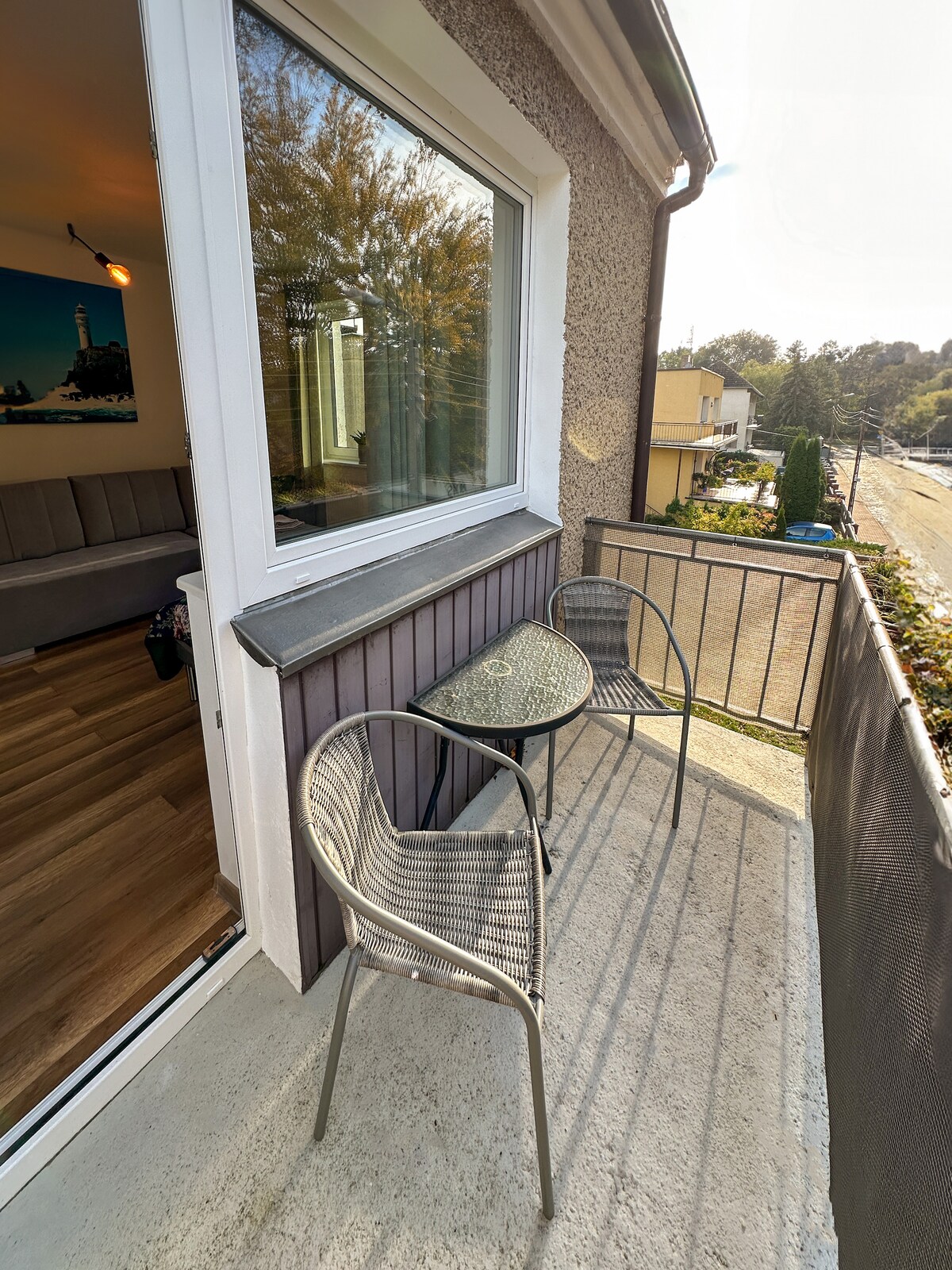 An outdoor balcony is depicted featuring a small round table surrounded by two lightweight chairs. A view of the surrounding landscape is visible through the large window, allowing natural light to brighten the space.
