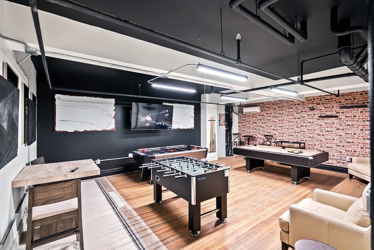 A spacious lounge area features a billiards table and a foosball table. Exposed brick walls provide an industrial backdrop, while modern seating options are arranged for socializing. Ambient lighting from overhead fixtures enhances the cozy atmosphere, highlighting entertainment options within the room.