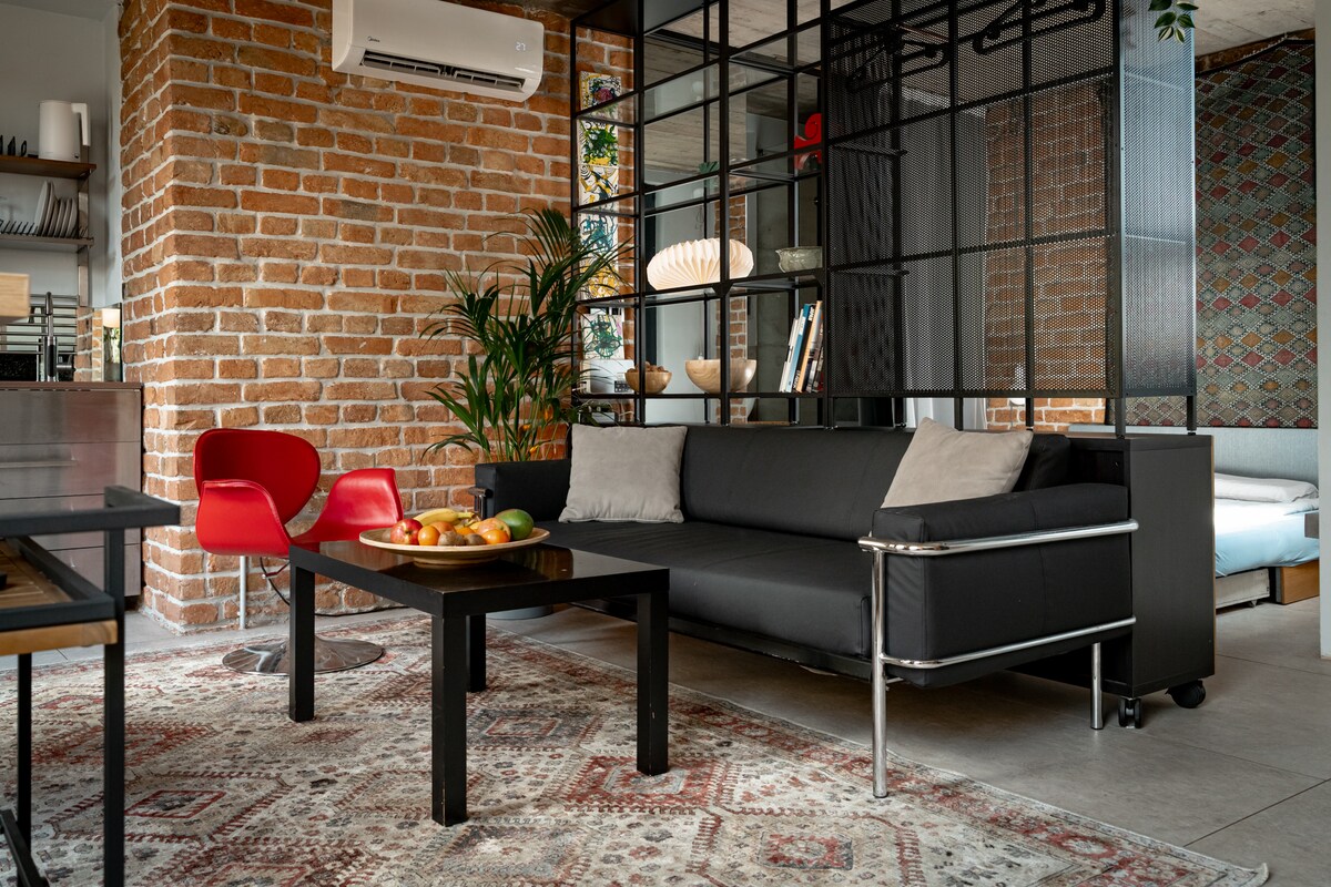 A modern living area features a black sofa with cushions, accented by a low coffee table displaying a bowl of fruit. Red chairs add a pop of color, while a brick wall and patterned backdrop provide texture. An air conditioning unit is installed overhead.