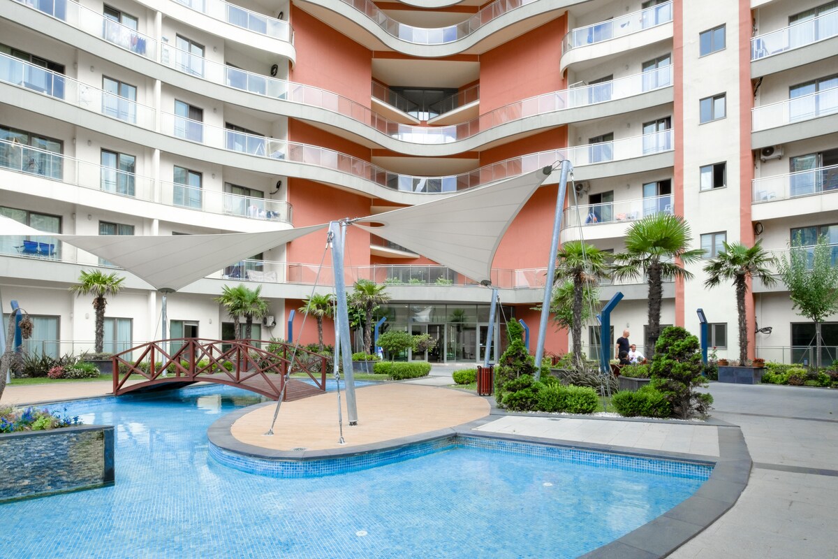 An outdoor area features a pool surrounded by landscaped greenery and stone pathways. A bridge crosses over the water, while shaded seating areas are provided beneath large awnings. The multi-story building forms the backdrop, showcasing a combination of red and white facades.