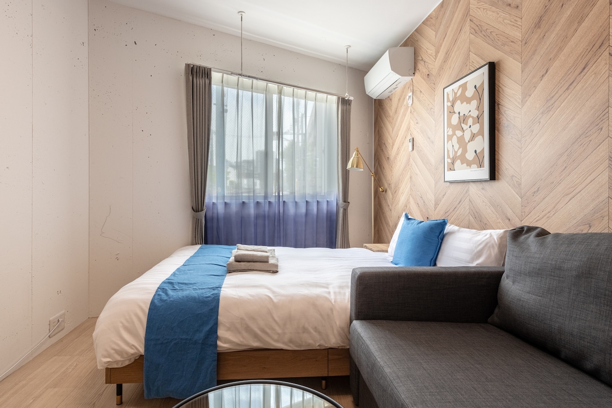 A serene sleeping area features a cozy bed with a blue accent blanket. A gray sofa is positioned nearby. Natural light filters through sheer curtains, illuminating the wooden wall with a geometric pattern. Decorative artwork adds a touch of design to the space.