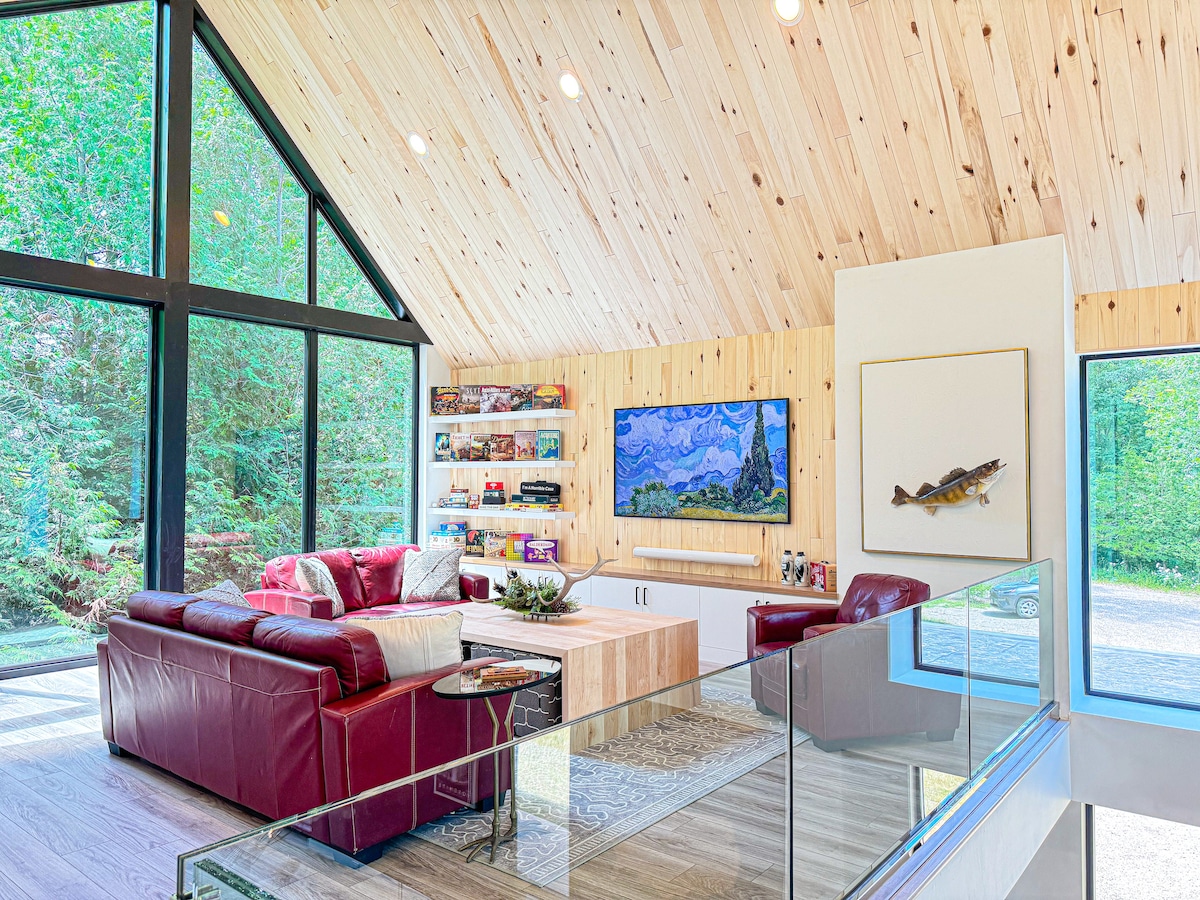 An inviting great room features a high, wooden ceiling that highlights natural textures. Large floor-to-ceiling windows provide ample sunlight, showcasing views of the outdoors. Comfortable seating is arranged around a central coffee table, while a television hangs above a sleek media console. A colorful piece of artwork adds character.