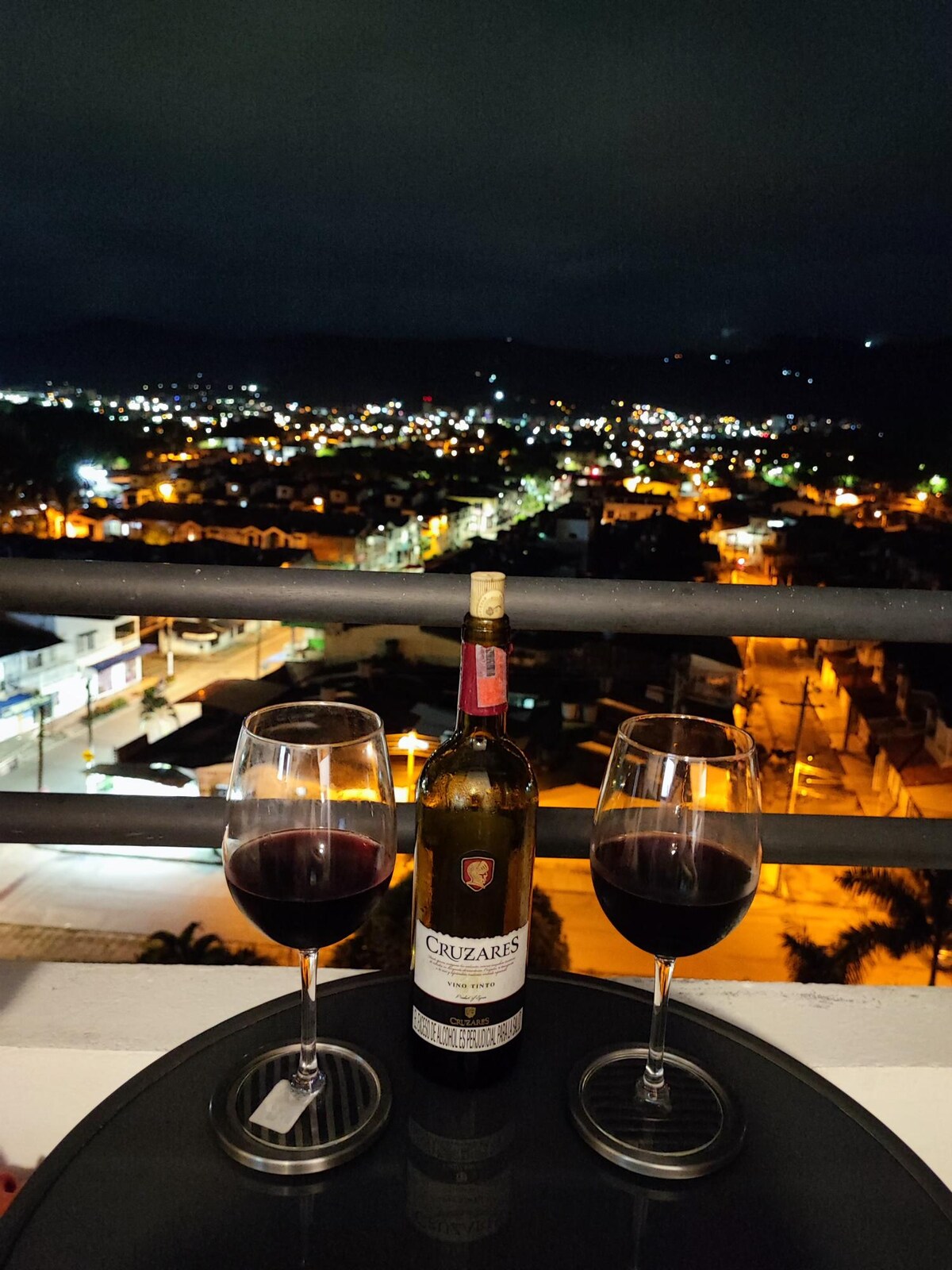 A cityscape at night is captured from a balcony, where two glasses of red wine are placed beside a bottle. The urban lights below shimmer, revealing roads and buildings illuminated against the dark sky, creating a cozy setting for relaxation.