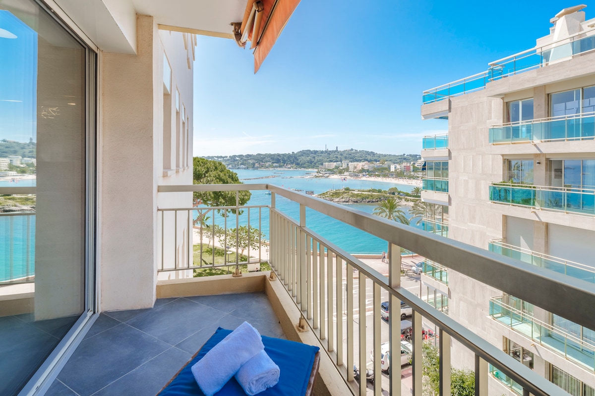 A spacious terrace features a comfortable lounge area with a sunbed and towels neatly arranged. A stunning view of the sea and the Cap d'Antibes can be seen in the background, while the bright blue sky enhances the overall ambiance.