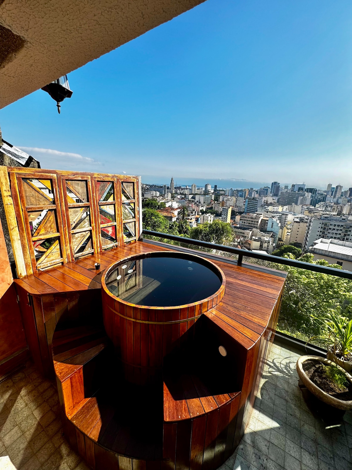 An outdoor balcony features a wooden Ofuro hot tub with a dark circular basin, surrounded by elegant wooden paneling. A scenic view of the city stretches across the background, complemented by clear blue skies. Potted greenery adds a natural touch to the space.