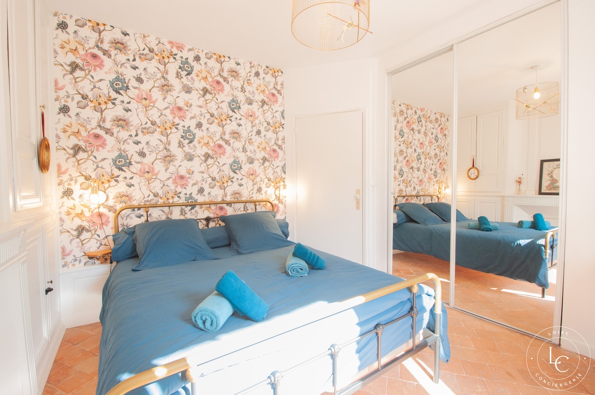 Chambre élégante avec lit queen size et décorée d'un papier peint floral coloré. Ambiance lumineuse grâce à la grande fenêtre et à l'armoire. Un bureau avec une chaise rose ajoute une touche de style et de fonctionnalité, parfait pour le télétravail.