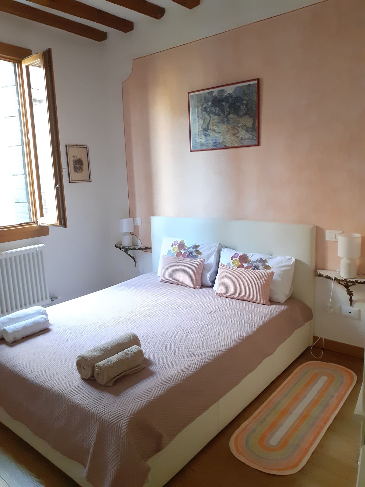A bright bedroom is featuring a spacious bed with a light-colored headboard. Decorative pillows add subtle color to the bedspread. Natural light enters through a window fitted with wooden frames, while a piece of art is displayed on the wall above. Soft towels are neatly arranged on the bed.