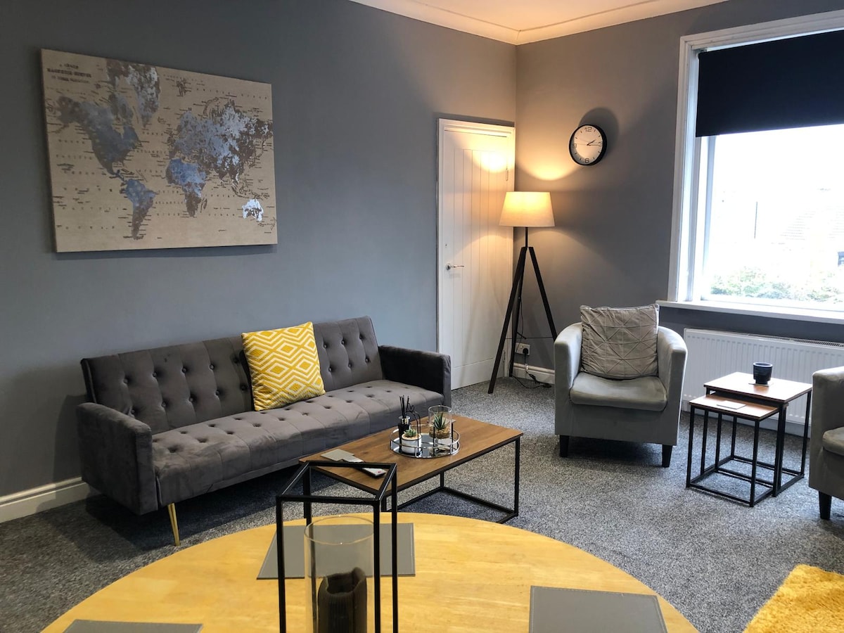 The living space features a modern style with a gray color palette. A cozy sofa, two armchairs, and a round coffee table are arranged invitingly. A world map artwork adorns the wall, and a floor lamp provides additional light. Natural light enters through a window, adorned with blackout blinds.