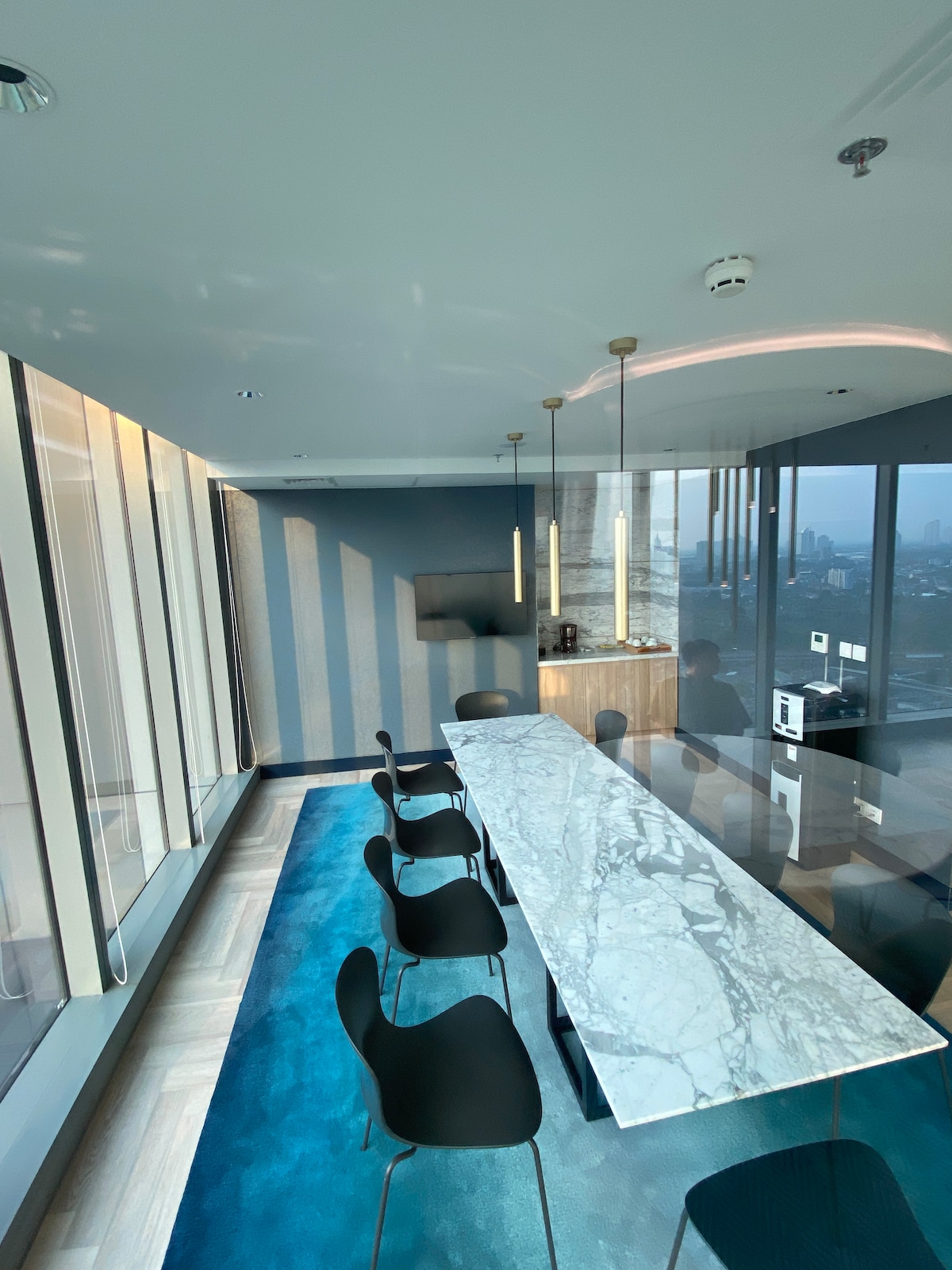 A modern dining area features a long marble table surrounded by several sleek black chairs. Large windows offer panoramic views of the city skyline, while soft natural light highlights the blue area rug beneath the table. Minimalist design elements create a clean and sophisticated atmosphere.